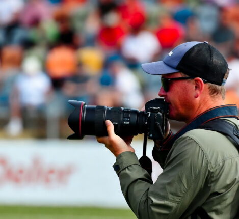 عکاس ورزشی در کنار زمین فوتبال در حال تمرکز بر جریان بازی برای پیش‌بینی لحظه حساس گل.