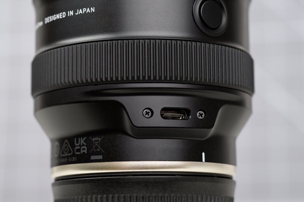 بررسی تخصصی لنز تامرون 25-200mm f/2.8-5.6 Di III VXD G2: سفر سبک و بیدغدغه 2 نمای نزدیک از حلقه زوم، سوئیچ قفل و پورت USB-C روی بدنه لنز تامرون 25-200mm.