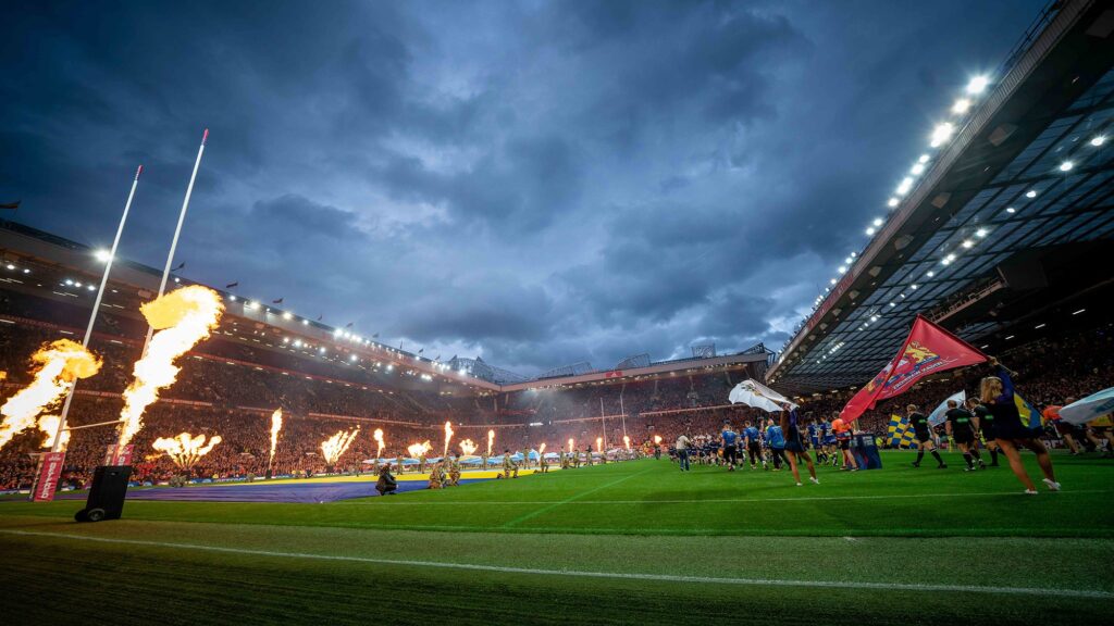 این عبارت به توصیف یک تصویر عریض از یک زمین راگبی قبل از شروع بازی اشاره دارد که شعله‌هایی در کنار زمین در حال بلند شدن هستند و ایجاد یک فضای جذاب و هیجان‌انگیز می‌کند.